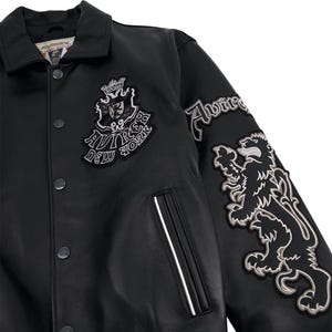 K&ouml;nnte beinhalten: Schwarze Lederjacke mit Kragen und Druckkn&ouml;pfen. Mit gestickten Aufn&auml;hern mit dem Text "AVIREX NEW YORK" und einer L&ouml;wen-Grafik. Ein wei&szlig;er Streifen akzentuiert die Tasche.