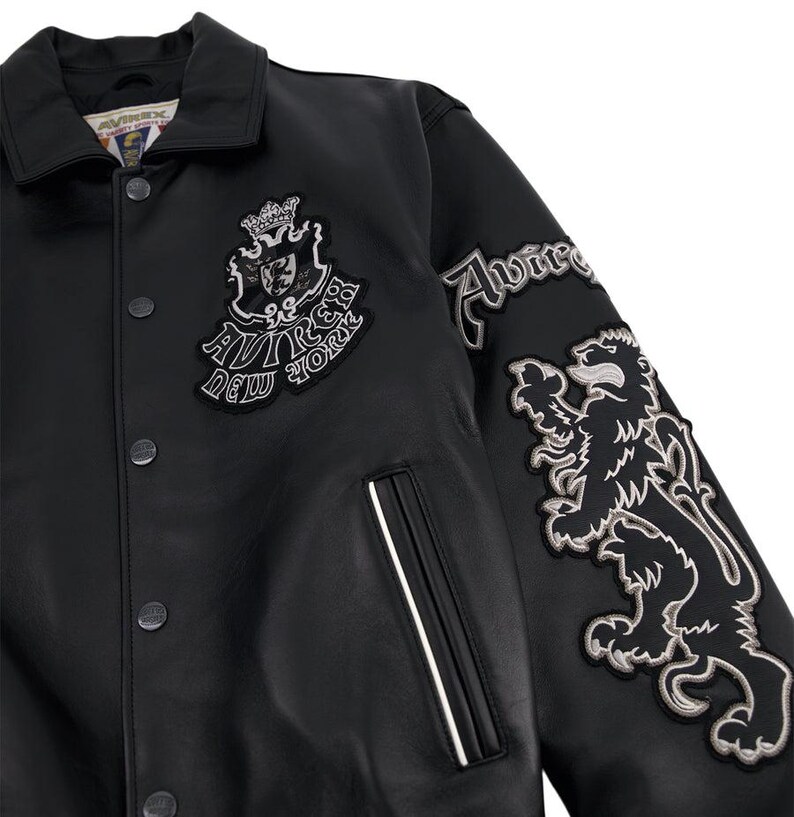 K&ouml;nnte beinhalten: Schwarze Lederjacke mit Kragen und Druckkn&ouml;pfen. Mit gestickten Aufn&auml;hern mit dem Text "AVIREX NEW YORK" und einer L&ouml;wen-Grafik. Ein wei&szlig;er Streifen akzentuiert die Tasche.