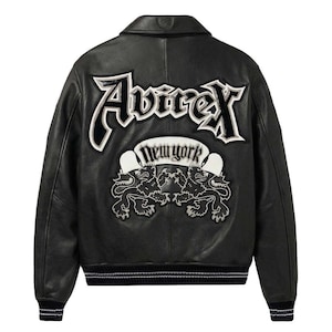 K&ouml;nnte beinhalten: Schwarze Lederjacke mit wei&szlig; gesticktem "Avirex New York" Logo und zwei L&ouml;wenwappen auf dem R&uuml;cken. Die Jacke hat einen klassischen Bomber-Stil mit geripptem Saum und B&uuml;ndchen.