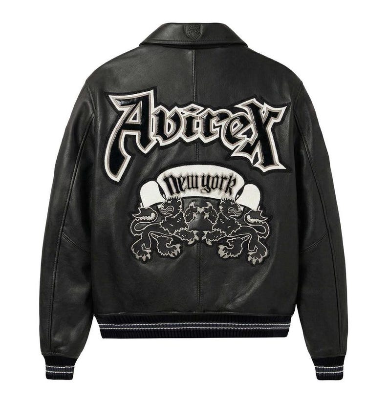 K&ouml;nnte beinhalten: Schwarze Lederjacke mit wei&szlig; gesticktem "Avirex New York" Logo und zwei L&ouml;wenwappen auf dem R&uuml;cken. Die Jacke hat einen klassischen Bomber-Stil mit geripptem Saum und B&uuml;ndchen.