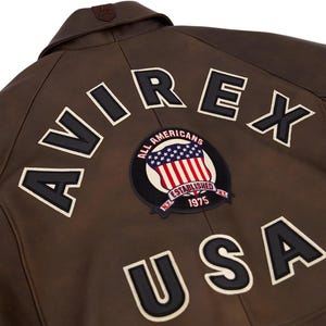 Op de afbeelding: Bruine leren jas met de woorden "AVIREX" en "USA" in grote, zwarte blokletters met witte contouren. Een ronde patch met de Amerikaanse vlag en de woorden "ALL AMERICANS ESTABLISHED 1975" is ook zichtbaar.