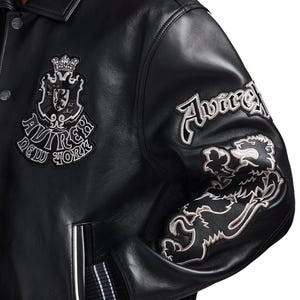 K&ouml;nnte beinhalten: Schwarze Lederjacke mit gestickten Aufn&auml;hern. Mit einem Wappen mit Krone und dem Text "AVIREX NEW YORK". Der rechte &Auml;rmel hat "Avirex" und einen L&ouml;wenaufn&auml;her. Die Jacke hat einen Knopfverschluss und gerippte B&uuml;ndchen.