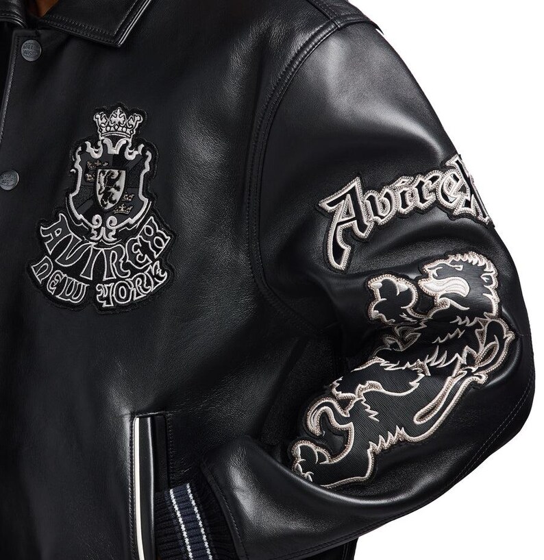 K&ouml;nnte beinhalten: Schwarze Lederjacke mit gestickten Aufn&auml;hern. Mit einem Wappen mit Krone und dem Text "AVIREX NEW YORK". Der rechte &Auml;rmel hat "Avirex" und einen L&ouml;wenaufn&auml;her. Die Jacke hat einen Knopfverschluss und gerippte B&uuml;ndchen.