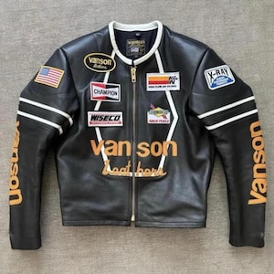 May include: Black leather motorcycle jacket with white stripes and patches. Features the brand name "Vanson" in orange on the sleeves and chest. Patches include "Champion", "Wiseco", and an American flag.