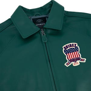 May include: A dark green leather jacket with a collar and a zipper closure. The jacket features a patch on the chest with the text "AVIREX" and an American flag design. The text "AUTHENTIC 1975" is also visible.