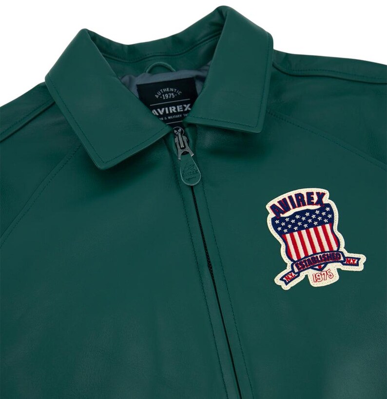 May include: A dark green leather jacket with a collar and a zipper closure. The jacket features a patch on the chest with the text "AVIREX" and an American flag design. The text "AUTHENTIC 1975" is also visible.