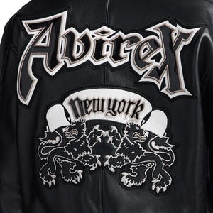 K&ouml;nnte beinhalten: Schwarze Lederjacke mit dem Wort "Avirex" in gro&szlig;en, wei&szlig; umrandeten Buchstaben auf dem R&uuml;cken. Darunter ein "New York" Banner und zwei L&ouml;wenembleme, wei&szlig; gestickt.