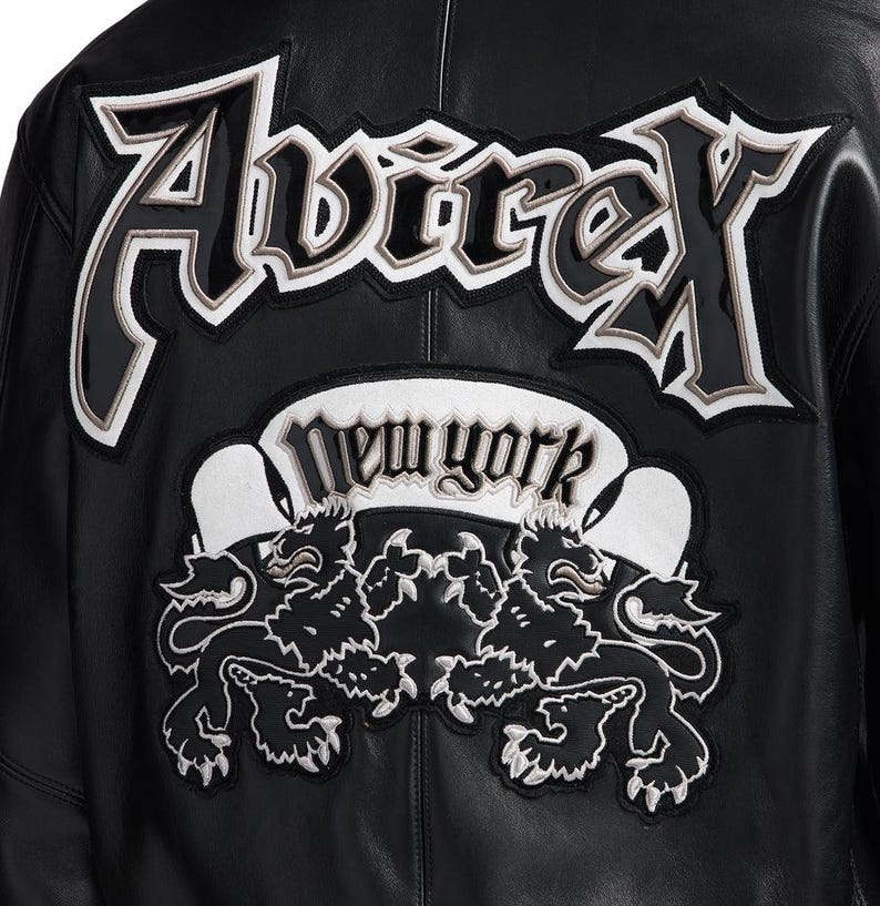 K&ouml;nnte beinhalten: Schwarze Lederjacke mit dem Wort "Avirex" in gro&szlig;en, wei&szlig; umrandeten Buchstaben auf dem R&uuml;cken. Darunter ein "New York" Banner und zwei L&ouml;wenembleme, wei&szlig; gestickt.