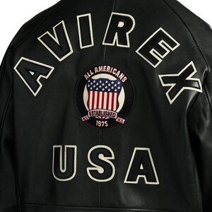 Op de afbeelding: Zwarte leren jas met witte geborduurde letters "AVIREX" en "USA". Een cirkelvormige patch toont de Amerikaanse vlag en de woorden "ALL AMERICANS ESTABLISHED 1975 N.Y."