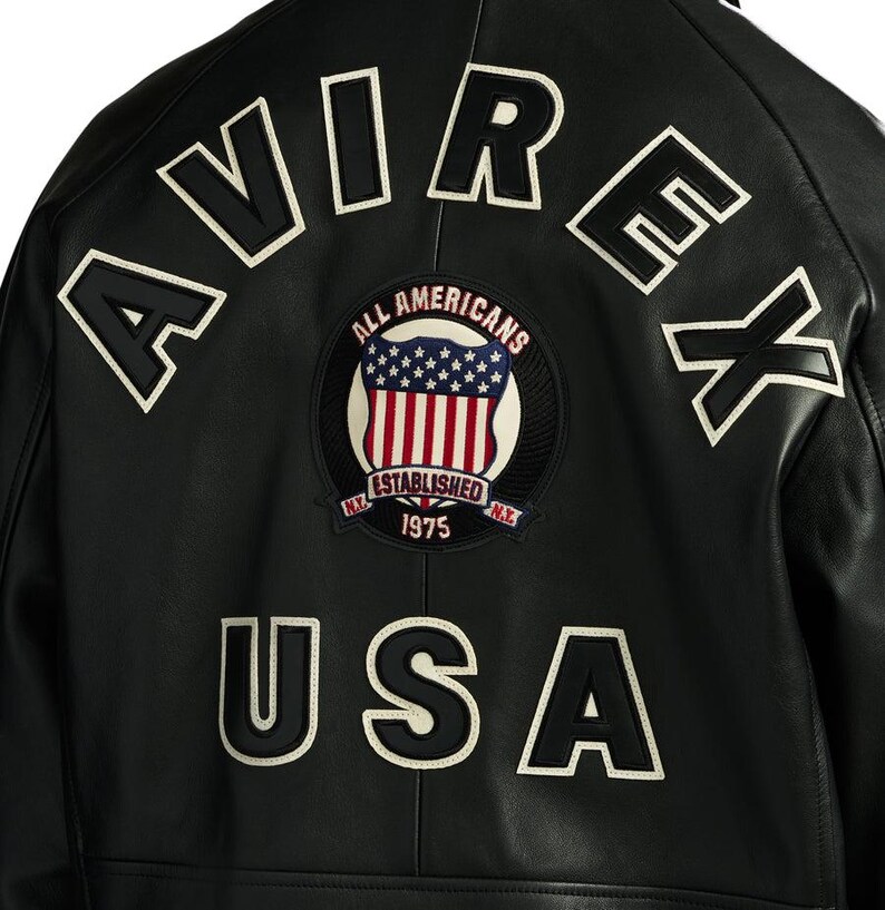 Op de afbeelding: Zwarte leren jas met witte geborduurde letters "AVIREX" en "USA". Een cirkelvormige patch toont de Amerikaanse vlag en de woorden "ALL AMERICANS ESTABLISHED 1975 N.Y."