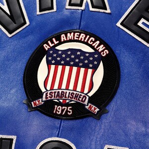 May include: A close-up of a blue leather jacket with white and black lettering. The jacket features a circular patch with the words "ALL AMERICANS" and "ESTABLISHED 1975 N.Y." The patch has a shield design with the American flag.