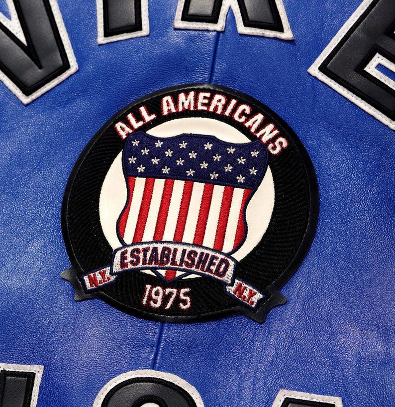 May include: A close-up of a blue leather jacket with white and black lettering. The jacket features a circular patch with the words "ALL AMERICANS" and "ESTABLISHED 1975 N.Y." The patch has a shield design with the American flag.