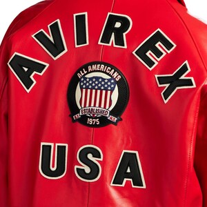 Peut inclure: Veste en cuir rouge avec le mot "AVIREX" en lettres blanches bord&eacute;es de noir sur les &eacute;paules. En dessous, un &eacute;cusson circulaire avec les mots "ALL AMERICANS" et "ESTABLISHED 1975" entoure un motif du drapeau am&eacute;ricain. Le mot "USA" est en dessous.