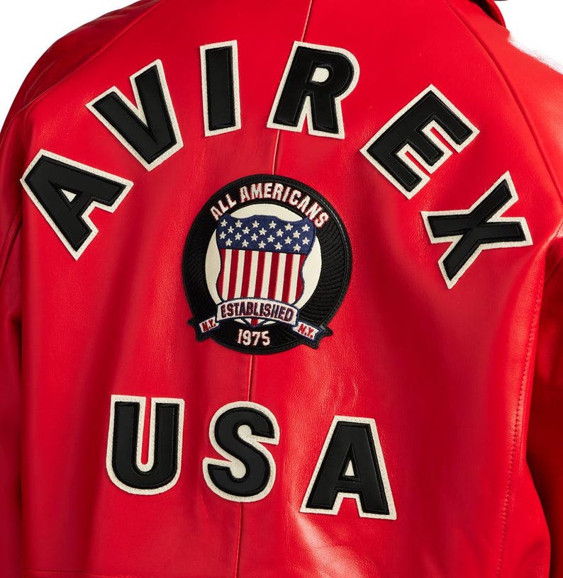 Peut inclure: Veste en cuir rouge avec le mot "AVIREX" en lettres blanches bord&eacute;es de noir sur les &eacute;paules. En dessous, un &eacute;cusson circulaire avec les mots "ALL AMERICANS" et "ESTABLISHED 1975" entoure un motif du drapeau am&eacute;ricain. Le mot "USA" est en dessous.