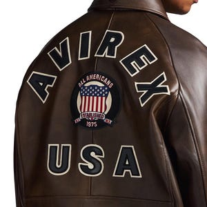 Op de afbeelding: Bruine leren jas met het woord "AVIREX" in witte letters met zwarte contouren op de rug. Daaronder een patch met de woorden "ALL AMERICANS" en "USA" in witte letters met zwarte contouren. De patch toont de Amerikaanse vlag.