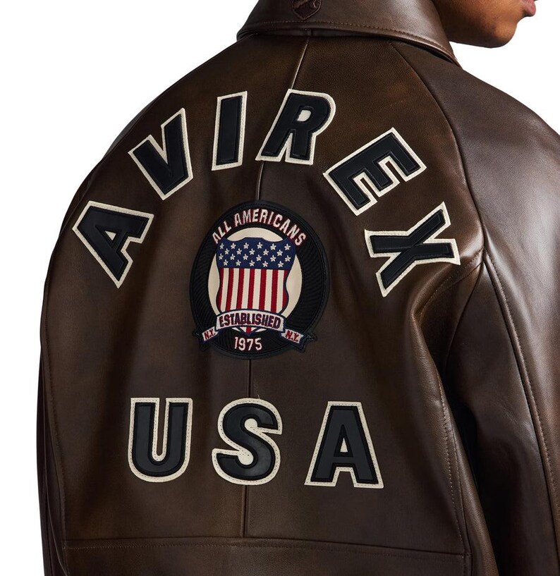 Op de afbeelding: Bruine leren jas met het woord "AVIREX" in witte letters met zwarte contouren op de rug. Daaronder een patch met de woorden "ALL AMERICANS" en "USA" in witte letters met zwarte contouren. De patch toont de Amerikaanse vlag.