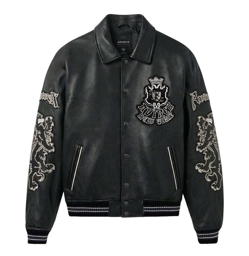 K&ouml;nnte beinhalten: Schwarze Leder-Collegejacke mit wei&szlig;en Akzenten. Mit gestickten L&ouml;wen und Textdetails auf den &Auml;rmeln und der Brust. Die Jacke hat einen Kragen, eine Knopfleiste und gerippte B&uuml;ndchen und Saum. Der Markenname "AVIREX NEW YORK" ist sichtbar.