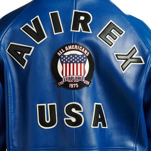 May include: A close-up of the back of a blue leather jacket. The jacket features white and black lettering that spells out "AVIREX" and "USA". A circular patch with the words "ALL AMERICANS" and "ESTABLISHED 1975" is also visible.