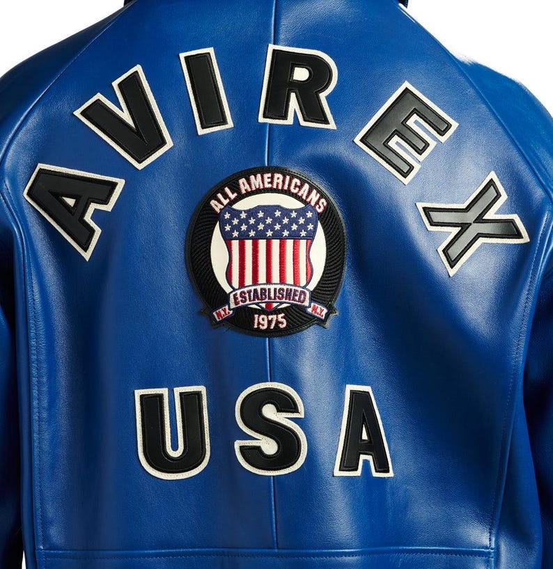 May include: A close-up of the back of a blue leather jacket. The jacket features white and black lettering that spells out "AVIREX" and "USA". A circular patch with the words "ALL AMERICANS" and "ESTABLISHED 1975" is also visible.