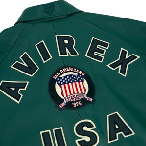 May include: A dark green leather jacket with white lettering spelling "AVIREX" and "USA". A circular patch features the American flag and the words "ALL AMERICANS ESTABLISHED NY 1975".