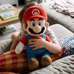 Peut inclure: Une peluche Mario, avec une casquette rouge ornée d'un "M" blanc, une moustache noire et une salopette bleue. La poupée est tenue par une personne sur un canapé gris avec des coussins colorés. La scène se déroule dans un salon lumineux.
