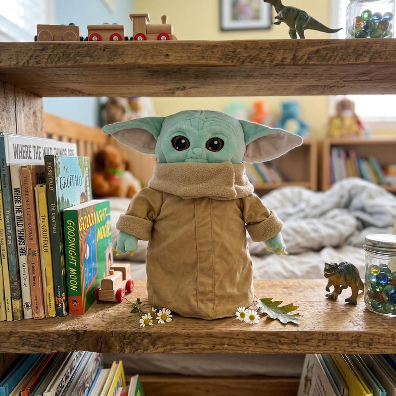 May include: A plush doll of a character with large ears and green skin, wearing a tan coat and a beige collar. The doll is on a wooden shelf with books and toys, including a wooden train set and a dinosaur figurine. The books include "Goodnight Moon" and "Where the Wild Things Are".