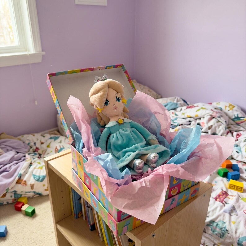 Puede incluir: Una mu&ntilde;eca de peluche con cabello rubio, un vestido azul y zapatos plateados se encuentra dentro de una caja decorativa llena de papel de seda rosa y azul. La caja est&aacute; colocada en una estanter&iacute;a de madera en un dormitorio.