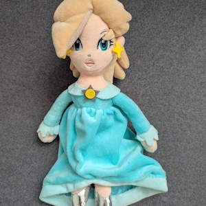 Puede incluir: Mu&ntilde;eca de peluche de un personaje con cabello rubio, ojos azules y un vestido azul claro. La mu&ntilde;eca tiene una corona plateada y pendientes en forma de estrella. El vestido de la mu&ntilde;eca tiene mangas largas y un cuello a juego. La mu&ntilde;eca lleva zapatos plateados.