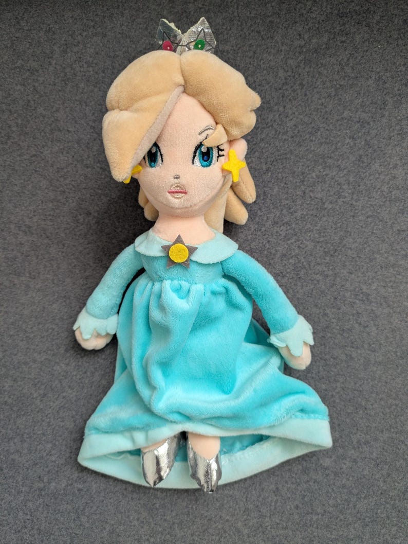 Puede incluir: Mu&ntilde;eca de peluche de un personaje con cabello rubio, ojos azules y un vestido azul claro. La mu&ntilde;eca tiene una corona plateada y pendientes en forma de estrella. El vestido de la mu&ntilde;eca tiene mangas largas y un cuello a juego. La mu&ntilde;eca lleva zapatos plateados.