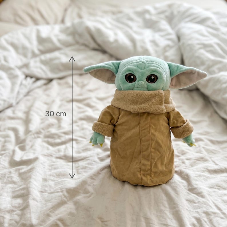 May include: A plush toy of a character with large ears, green skin, and big black eyes. The toy is wearing a tan coat and has a beige collar. The toy measures approximately 11.8 inches in height.