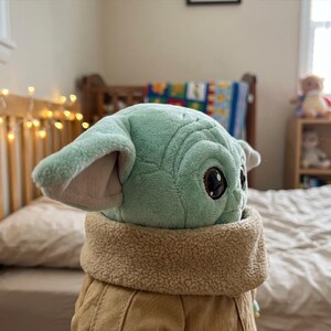 May include: A plush toy of a character with large ears and big eyes, wearing a tan coat with a fuzzy collar. The toy is light green and tan. The toy is sitting on a bed with a white comforter.