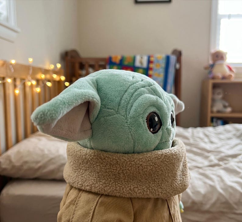 May include: A plush toy of a character with large ears and big eyes, wearing a tan coat with a fuzzy collar. The toy is light green and tan. The toy is sitting on a bed with a white comforter.