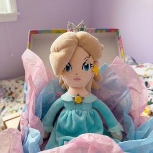 Puede incluir: Mu&ntilde;eca de peluche de un personaje con cabello rubio, ojos azules y un vestido azul claro, dentro de una caja decorativa con papel de seda rosa y azul. La mu&ntilde;eca tiene una corona y pendientes en forma de estrella.