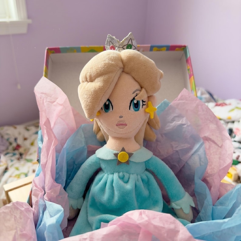 Puede incluir: Mu&ntilde;eca de peluche de un personaje con cabello rubio, ojos azules y un vestido azul claro, dentro de una caja decorativa con papel de seda rosa y azul. La mu&ntilde;eca tiene una corona y pendientes en forma de estrella.