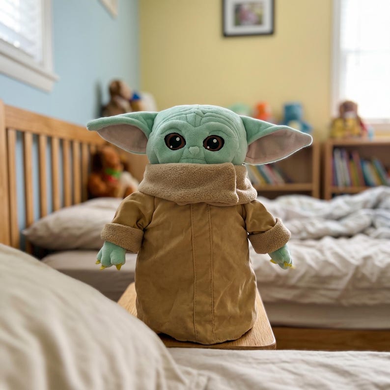 May include: A plush toy of a character with large eyes, green skin, and large ears. The character is wearing a tan coat and a beige scarf. The toy is sitting on a wooden surface in a bedroom setting.