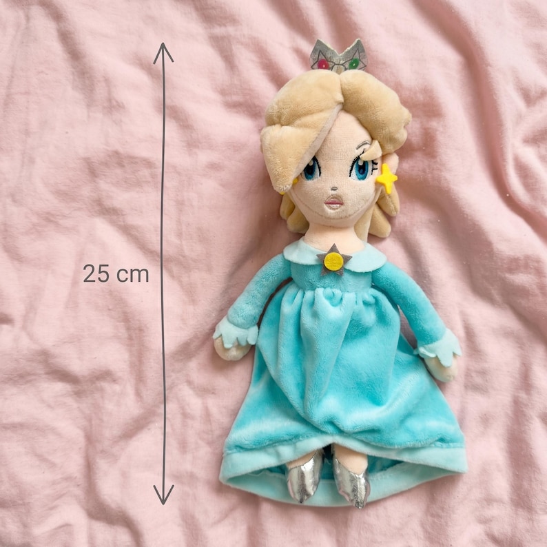 Puede incluir: Mu&ntilde;eca de peluche de un personaje con cabello rubio, ojos azules y una corona plateada. La mu&ntilde;eca lleva un vestido azul claro y zapatos plateados. La mu&ntilde;eca mide 25 cm de altura.