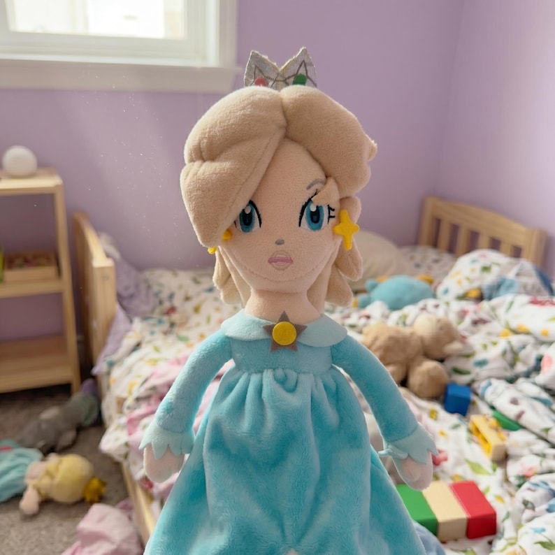 Puede incluir: Mu&ntilde;eca de peluche de un personaje con cabello rubio, ojos azules y un vestido azul claro. La mu&ntilde;eca tiene una corona y pendientes en forma de estrella. La mu&ntilde;eca est&aacute; en el dormitorio de un ni&ntilde;o con una cama y otros juguetes.