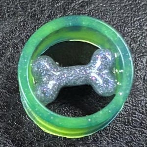 May include: A green and yellow ring with a glittery, gray dog bone-shaped charm in the center. The ring has a translucent appearance, and the bone charm sparkles with iridescent glitter.