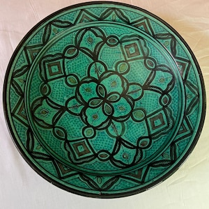 May include: A round, decorative ceramic plate with a teal and black geometric pattern. The plate features a central floral design surrounded by diamond and oval shapes. The rim is black, and the plate has a glossy finish.