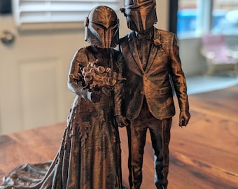 May include: A 3D printed bronze-colored figurine of a couple standing side by side. The figure on the left wears a long gown and holds a bouquet, while the figure on the right wears a suit. Both figures have helmets resembling a Mandalorian mask.