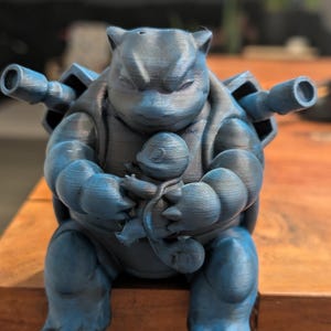 May include: A 3D printed blue figurine of a large turtle-like creature with two cannons on its back, holding a smaller, similar-looking creature in its arms. The figure has a textured surface and is colored in shades of dark and light blue.