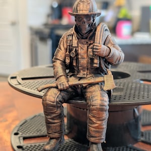 May include: A bronze-coloured figurine of a firefighter with a skull face, wearing a helmet and protective gear, holding a coiled hose over one shoulder and an axe across the lap. The figure is seated, featuring detailed textures on the uniform.