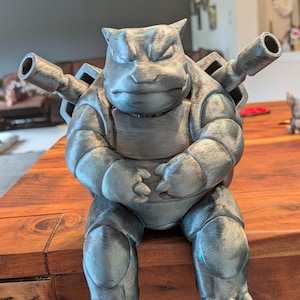 May include: A metallic silver-colored 3D printed figurine of the creature Blastoise, featuring a large shell, two prominent cannons extending from the back, a rounded body, and clawed feet, rendered with a textured, layered finish throughout.