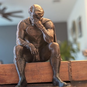 May include: A bronze-colored 3D printed figurine of a superhero character sitting with a head bowed and a hand covering the face. The figure wears a suit with a prominent chest emblem, a cape, and boots, featuring a textured surface finish throughout.