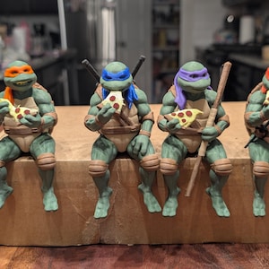 Puede incluir: Cuatro figuras de las Tortugas Ninja con piel verde y caparazones de color tostado. Cada figura lleva una máscara de un color diferente: naranja, azul, morado y rojo. Sostienen porciones de pizza y una figura sostiene un bastón de madera.
