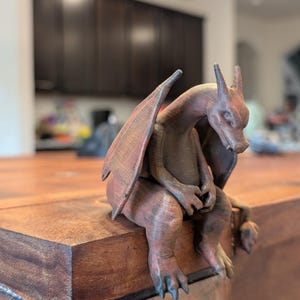 Puede incluir: Una figura de dragón impresa en 3D con un acabado texturizado de color marrón rojizo. La criatura se muestra en una postura sentada y encorvada con las alas parcialmente extendidas, garras afiladas en los pies y una larga cola colgando.