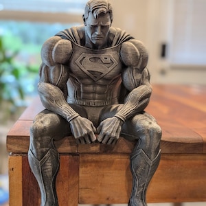 May include: A grey, stone-textured figurine of a muscular superhero sitting with a downward gaze. The figure features a prominent chest emblem, a cape draped over the shoulders, and detailed musculature on the arms, chest, and legs.