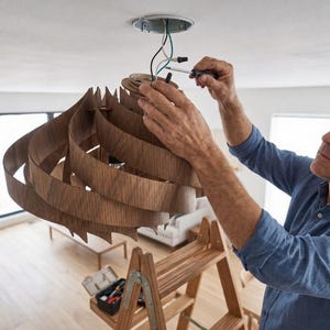 May include: A person fitting a wooden pendant light. The light fitting has a layered, swirling design with a natural wood finish. The person is using a screwdriver to connect the wires to the ceiling fixture. A wooden ladder and toolbox are also visible.