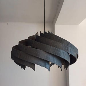 May include: A dark grey, spiral-shaped pendant light. The lamp hangs from a black cord and has layered, curved wooden slats with vertical grooves. The design is modern and architectural, suitable for a living space.