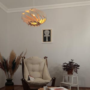 May include: A decorative wooden pendant light with a layered design hangs from the ceiling. The light fixture is made of light-coloured wood and emits a warm glow. The room also features a beige armchair, a framed artwork, and a potted plant.